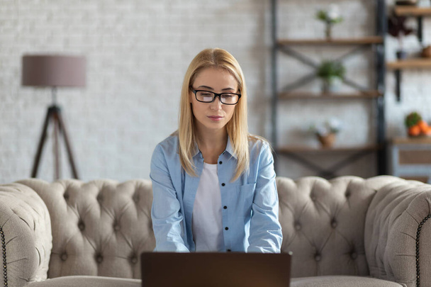 Serious beautiful young woman in eyeglasses working using laptop while working indoors - Fotó, kép