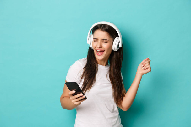 Portret van modern mooi meisje in wit t-shirt, luistermuziek in draadloze hoofdtelefoon, mobiele telefoon vasthouden en dansen, over een blauwe achtergrond staan - Foto, afbeelding