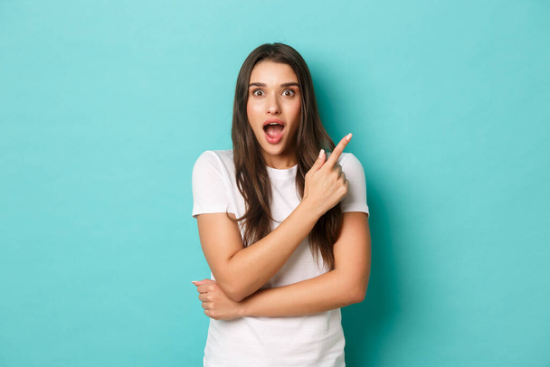 Cheerful brunette girl in white t-shirt, smiling excited and pointing fingers at upper right corner, showing amazing logo, standing over blue background - Fotografie, Obrázek