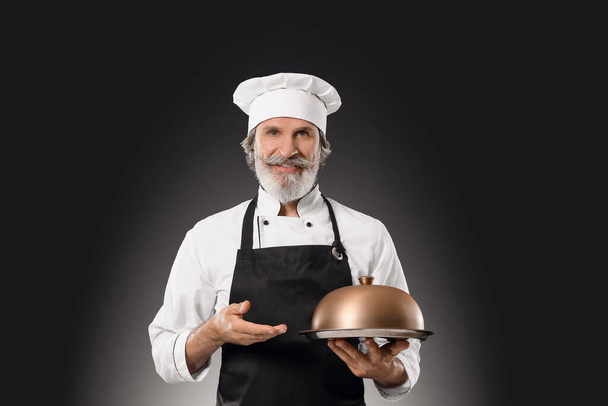 Chef macho maduro con bandeja y cloche sobre fondo oscuro - Foto, imagen