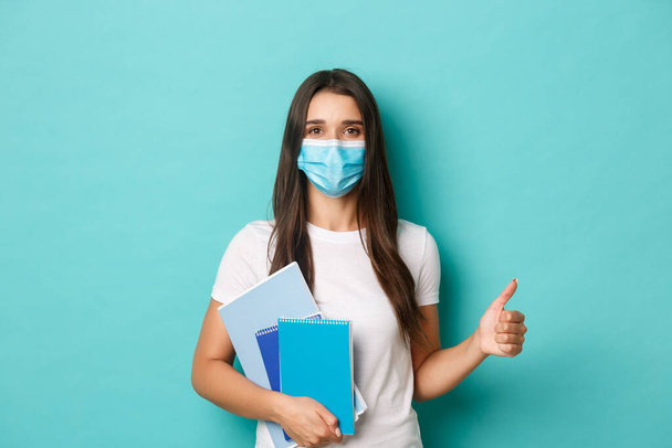 Concetto di coronavirus, salute e distanza sociale. Ritratto di studentessa frequentare le lezioni in maschera medica, mostrando pollici in alto in approvazione, tenendo quaderni, in piedi su sfondo blu - Foto, immagini