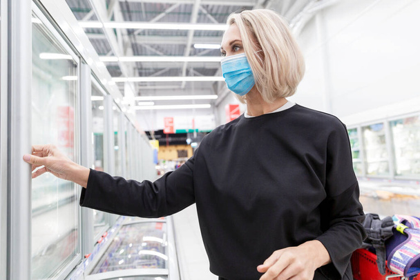 Young blond woman in mask in a supermarket. Coronavirus pandemic. - Фото, изображение