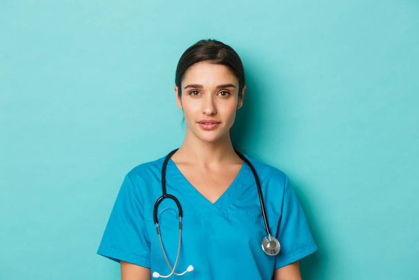 Concept of covid-19 and quarantine concept. Close-up of young confident female doctor with stethoscope, wearing scrubs, standing over blue background - Photo, Image