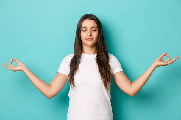 Image of peaceful pretty girl in white t-shirt, close eyes and spread hands sideways, meditating, practice yoga over blue background - Foto, Bild
