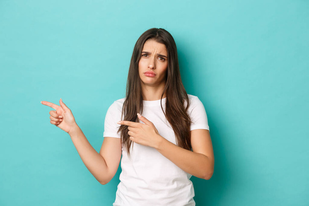 Portrait of sad and disappointed brunette girl in white t-shirt, frowning upset, pointing fingers left at copy space, standing over blue background - Foto, afbeelding