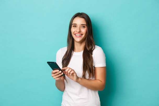 Portret van een moderne jonge vrouw in wit t-shirt, met behulp van een mobiele telefoon voor online winkelen of netwerken, staande over een blauwe achtergrond - Foto, afbeelding