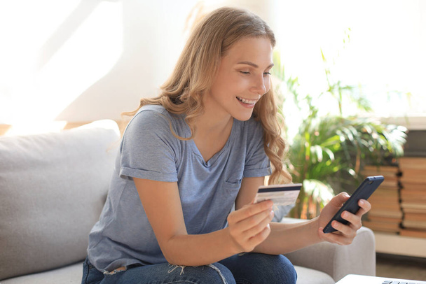 Happy natural blonde using credit card and mobile phone in living room - Photo, Image