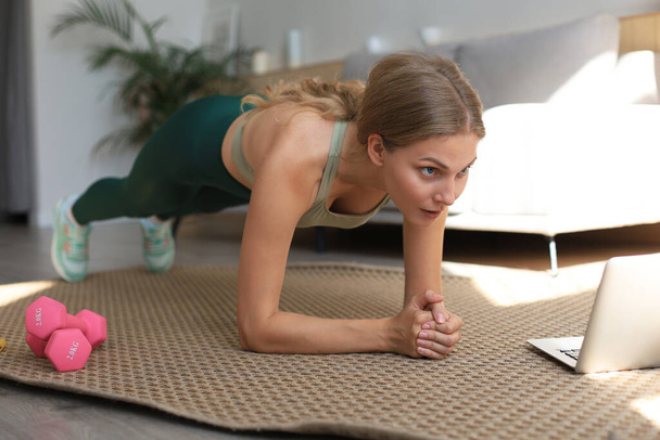 Fitness woman doing plank and watching online tutorials on laptop, training in living room. Stay at home activities. - Foto, imagen