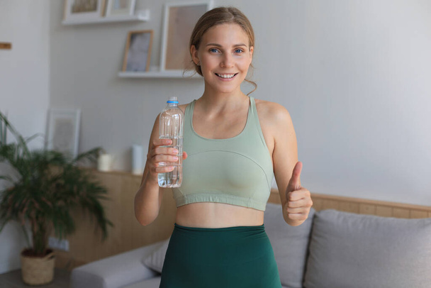Young beautiful woman standing with dumbbell and drink water after exercising looking camera. Attractive female bodybuilder working out. Fitness and healthy lifestyle concept. - Foto, imagen