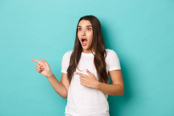 Excited and amazed beautiful girl in white t-shirt, pointing fingers and looking left at awesome advertisement, standing over blue background - Photo, Image