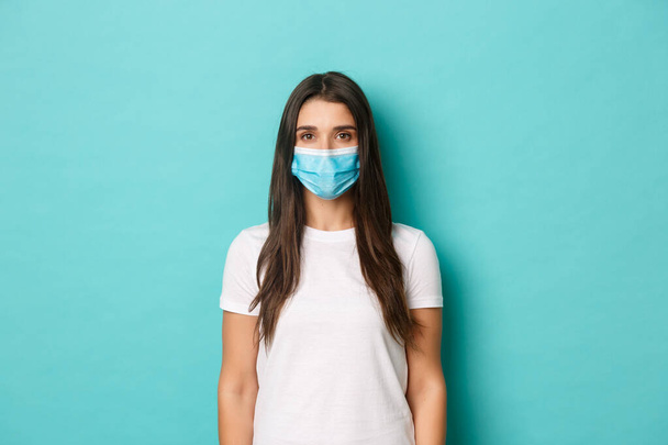 Koncepcja pandemii, covid-19 i dystansu społecznego. Młoda zdeterminowana kobieta w białej koszulce w masce medycznej, patrząca na aparat poważnie, stojąca nad niebieskim tłem - Zdjęcie, obraz