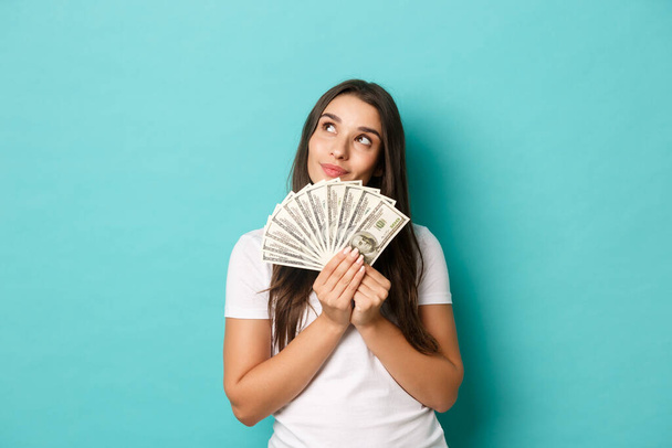Portrait of attractive brunette woman in white t-shirt, dreaming about shopping, holding money, standing over blue background - Фото, зображення