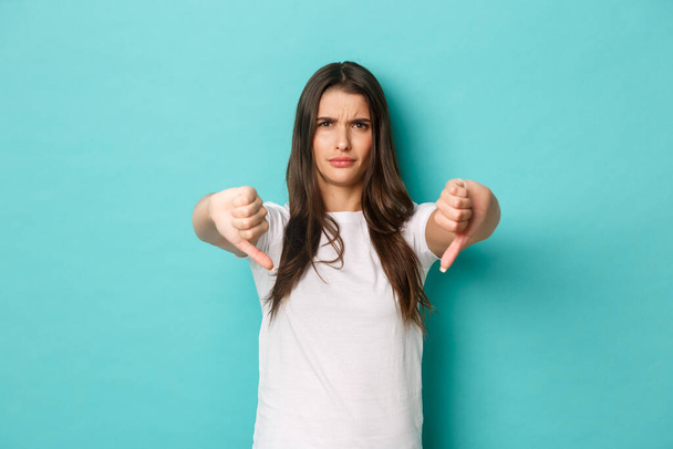 Desapontada mulher atraente em t-shirt branca, expressar sua discordância, mostrando polegares para baixo e olhando cético, de pé sobre fundo azul - Foto, Imagem