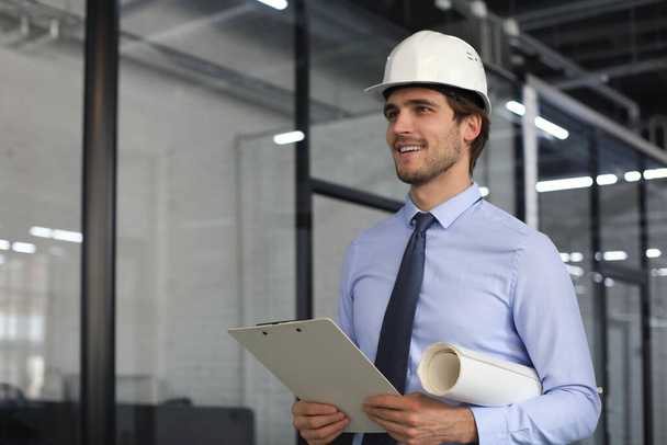 Confident young business man in hardhat holding blueprint and looking away while standing indoors - Fotoğraf, Görsel