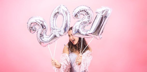 Aranyos fiatal lány mosollyal ünnepi ruhában pózol rózsaszín stúdió háttér és gazdaság ezüst lufik 2021 számok - Fotó, kép