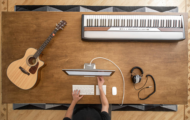 Male hands at a computer on a wooden table with an acoustic guitar, musical keys and studio headphones top view. - Foto, Bild