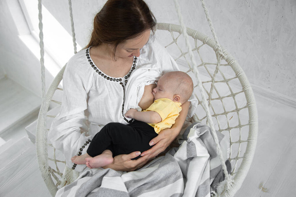 Mama geeft haar pasgeboren baby borstvoeding in een witte hangstoel op een lichte achtergrond. Borstvoeding concept, close-up, bovenaanzicht. - Foto, afbeelding
