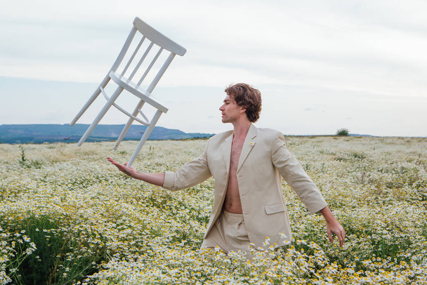 Tall handsome man standing in the middle of camomile flowers field and balancing with white chair on his palm - Фото, изображение