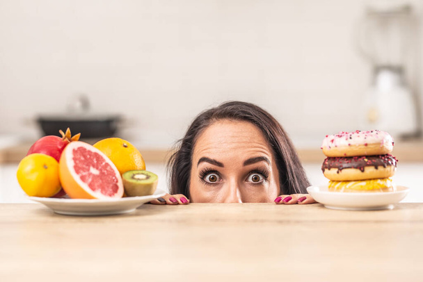 Választási lehetőség egy nő számára, aki az asztal fölött kukucskál, ahol gyümölcsökkel és fánkokkal teli tányér próbálja megragadni a figyelmét.. - Fotó, kép