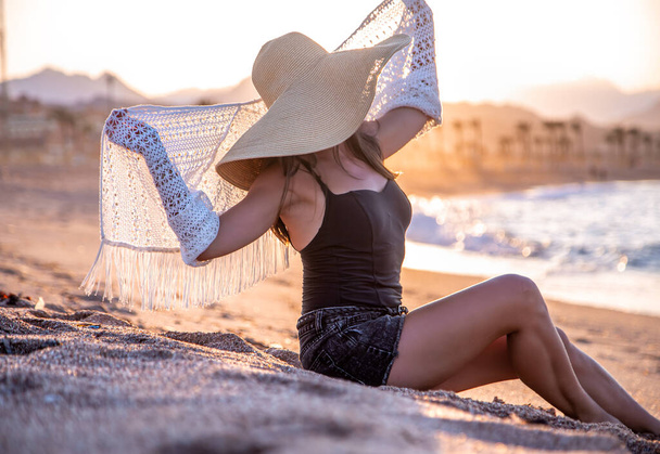 Beautiful boho model in a swimsuit with a white Cape with a hat on her head poses on the beach at sunset - Zdjęcie, obraz