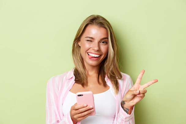 Close-up of attractive happy girl with blond hair, showing tongue and winking silly, make peace sign, using mobile phone, standing over green background - Fotografie, Obrázek