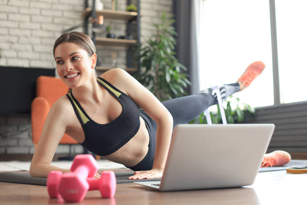 Fitness gyönyörű karcsú nő csinál oldalsó deszka egy rugalmas zenekar és néz online tutorials laptopon, edzés a nappaliban. Maradj otthon!. - Fotó, kép