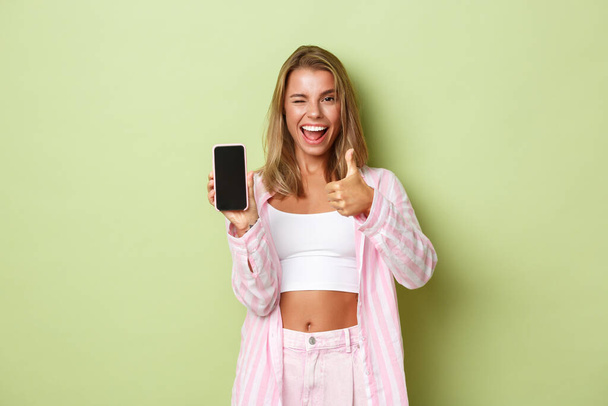 Красивая женщина в розовой рубашке, показывающая что-то хорошее на экране смартфона, поднимает вверх большие пальцы в одобрении и улыбается, стоя на зеленом фоне - Фото, изображение