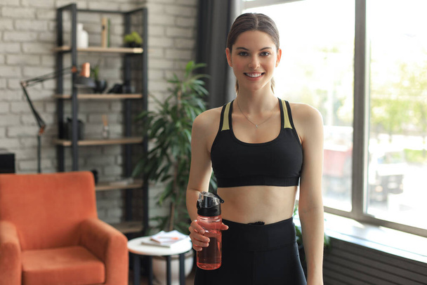 Young beautiful woman standing with dumbbell and drink water after exercising looking camera. Attractive female bodybuilder working out. Fitness and healthy lifestyle concept - 写真・画像