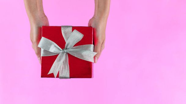Manos femeninas sosteniendo caja de regalo envuelta en papel rojo con cinta sobre fondo de color rosa. Vista superior. Copia espacio para texto. Concepto de vacaciones. - Foto, Imagen