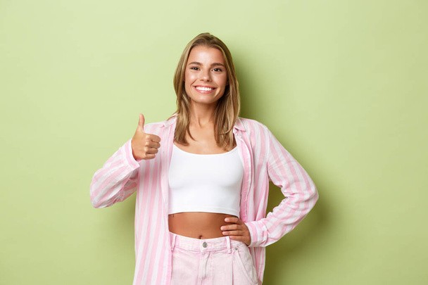 Beeld van zelfverzekerde moderne vrouw met blond kort kapsel, het dragen van roze shirt en jeans, met duimen-up in goedkeuring, als en aanbevelen iets goeds, groene achtergrond - Foto, afbeelding