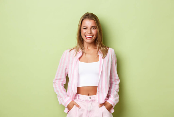 Портрет дерзкой современной женщины с короткими светлыми волосами, одетой в розовую одежду, показывающей язык и улыбающейся с идеальными белыми зубами, уверенно стоящей на зеленом фоне - Фото, изображение