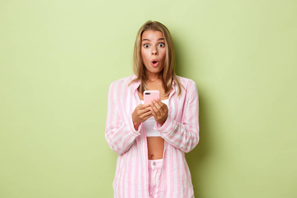 Portrait of blond woman in stylish pink outfit, looking surprised after reading something on mobile phone app, standing over green background - Foto, imagen