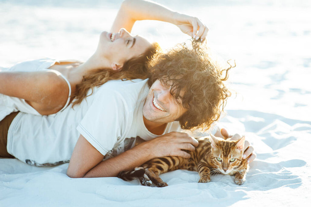 Cheerful young couple having fun on the beach with their bengal cat - Fotó, kép
