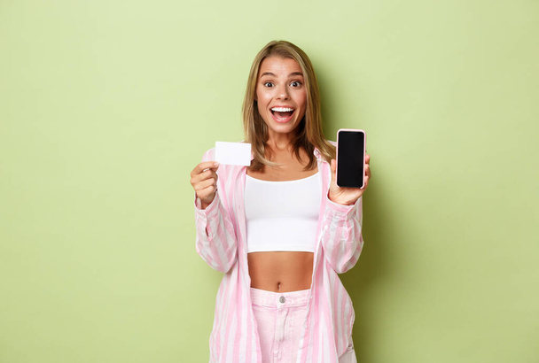 Image of stylish blond woman in pink shirt and jeans, showing smarpthone screen and credit card, recommending something online, standing over green background - Foto, afbeelding