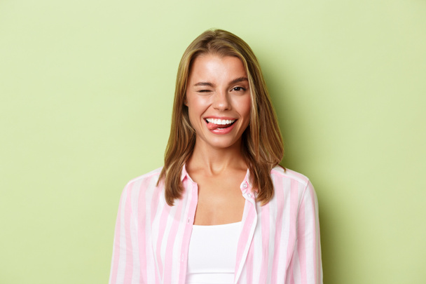 自信満々の笑顔の女性とブロンド髪,ショー舌とウィンクコケティッシュ,立っている上の緑の背景にピンクのシャツ - 写真・画像