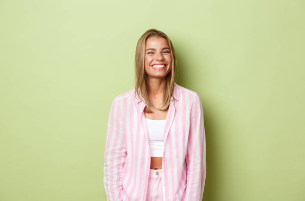 Imagen de una mujer rubia alegre con una sonrisa blanca perfecta, de pie sobre un fondo verde feliz, con un atuendo rosa - Foto, imagen