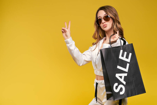 Beautiful smiling young girl in a sports suit and suglasses with a shopping bag on a yellow background. - Foto, Imagem