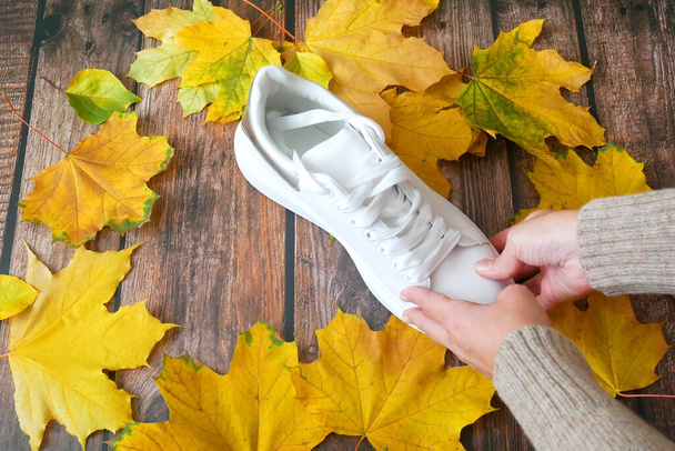 hand puts modern white sneakers and on wooden background with autumn leaves, autumn mood concept, autumn sales, flay lay - Photo, Image