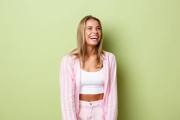 Portret van gelukkige mooie vrouw met kort blond haar, begint te lachen en kijkt naar de rechter bovenhoek, glimlachend gelukkig, staand in roze shirt en jeans over groene achtergrond - Foto, afbeelding