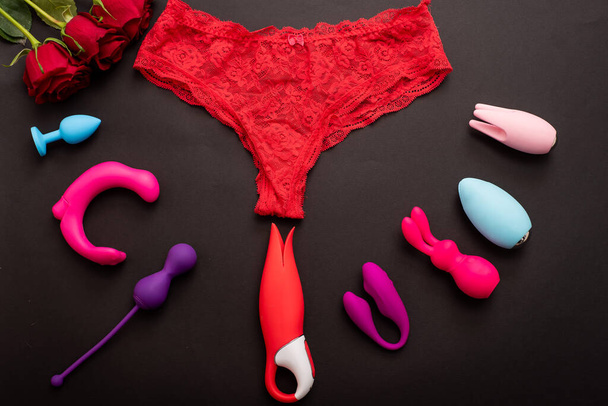 Top view of womens lace panties red roses and different sex toys. Collection of multicolored vibrators on a black background. - Foto, imagen