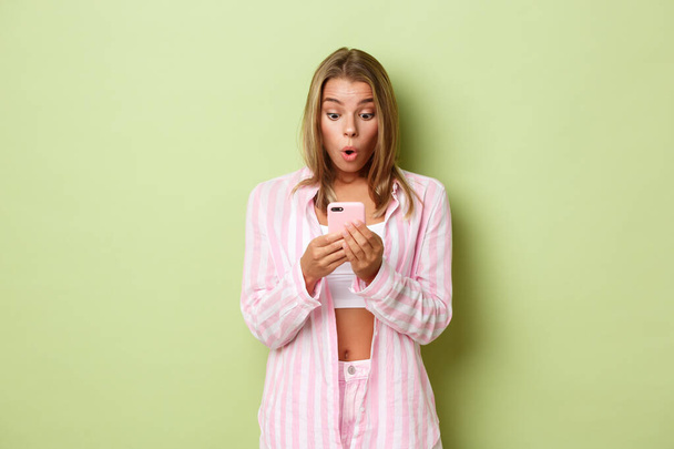 Portret zadziwionej blondynki ubranej w różową koszulę, zaskoczonej ekranem telefonu komórkowego, reagującej na wiadomość lub powiadomienie, stojącej nad zielonym tłem - Zdjęcie, obraz