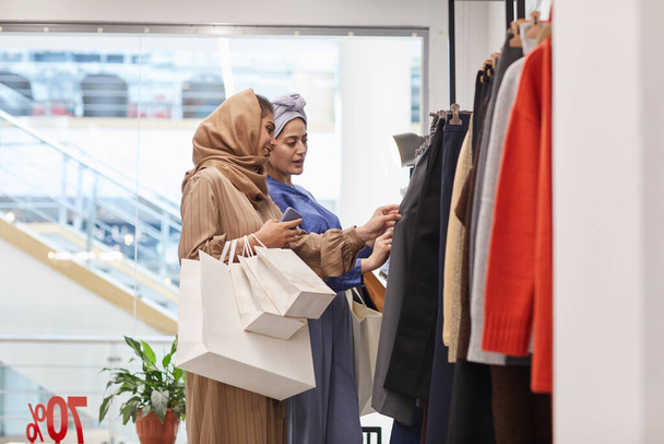 Portrait latéral de deux jeunes femmes du Moyen-Orient choisissant des vêtements tout en appréciant le shopping dans le centre commercial, espace de copie - Photo, image