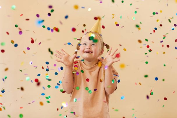 Waist up portrait of excited girl with down syndrome smiling happily while standing under confetti shower in studio, copy space - Fotografie, Obrázek
