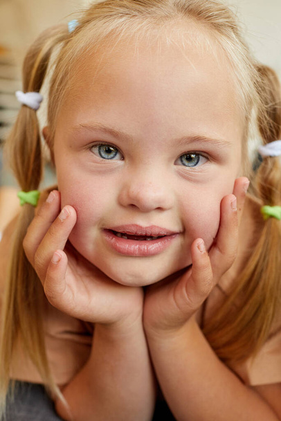 Vertikales Porträt eines süßen Mädchens mit Down-Syndrom, das lächelt und in die Kamera schaut, während es zu Hause auf dem Sofa liegt - Foto, Bild