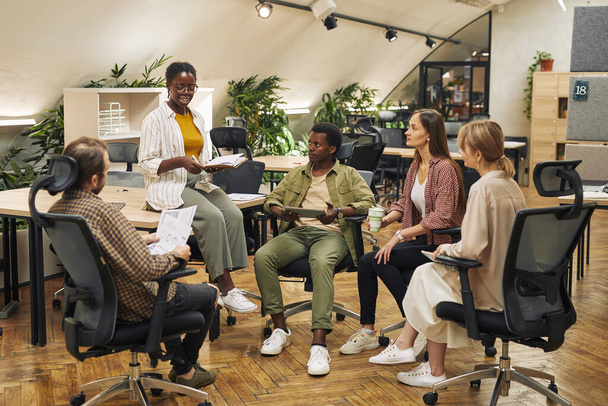 Pełnowymiarowy portret wieloetnicznej grupy młodych ludzi dyskutującej o projekcie pracy, siedzącej w kręgu w nowoczesnym biurze i słuchającej kierowniczki - Zdjęcie, obraz
