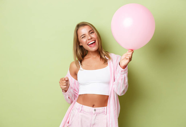 Obraz pięknej szczęśliwej kobiety z blond krótkimi włosami, zabawy na imprezie, świętowanie coś z różowym balonem, stojąc nad zielonym tle - Zdjęcie, obraz