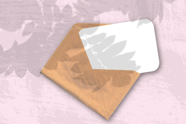 Конверт с текстурированной белой бумагой на розовом фоне стола. Наложение макета на тени растений. Естественный свет отбрасывает тени от ветки Роуэн - Фото, изображение