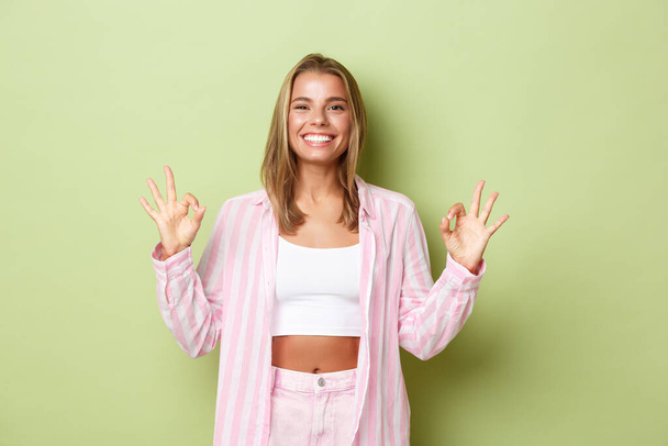 Portrait of attractive stylish woman with blond short hairstyle, wearing pink shirt with jeans, showing okay sign in approval, recommend something good, standing over green background - Photo, Image