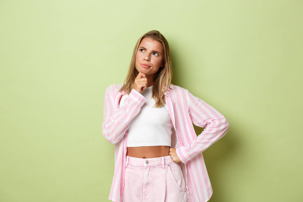 Εικόνα σκεπτόμενης νεαρής γυναίκας με ροζ στολή, που κοιτάζει την πάνω αριστερή γωνία και σκέφτεται, κάνει δύσκολη επιλογή, στέκεται πάνω από το πράσινο φόντο - Φωτογραφία, εικόνα