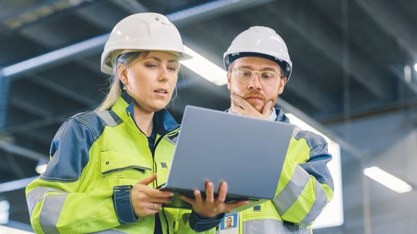 Low Angle Shot of Male and Female Industrial Engineers Work on a Manufacturing Plant, They Discuss Project while Using Laptop.In the Background Industrial Manufacturing Plant. - 写真・画像